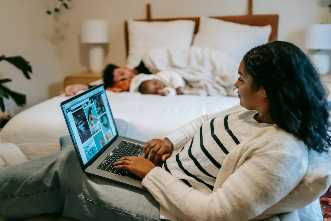 Mom on computer doing work and baby on bed coddle 