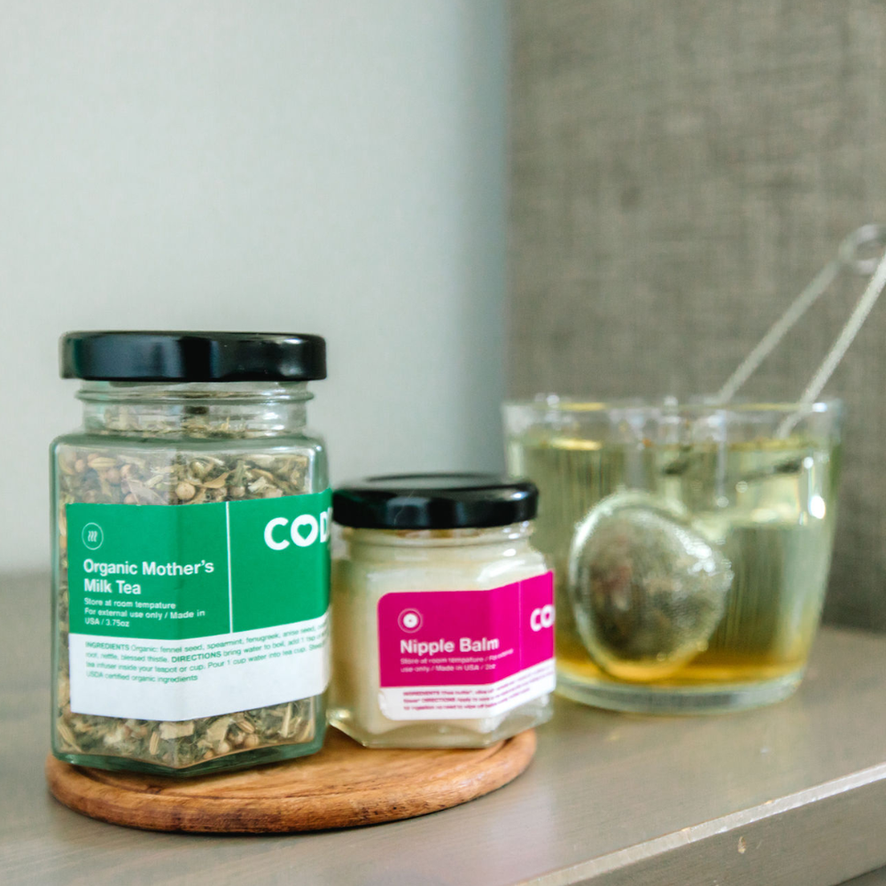 Jars of CODDLE mother's milk tea and nipple balm on a surface with a glass of tea in the background.
