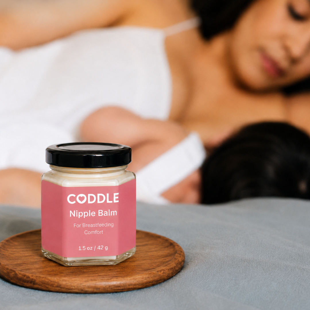 Coddle Nipple Balm sits on a wooden coaster on a bed, with a blurred image of woman in background breastfeeding.
