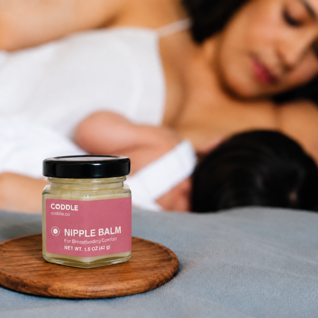 Coddle Nipple Balm sits on a wooden coaster on a bed, with a blurred image of woman in background breastfeeding.