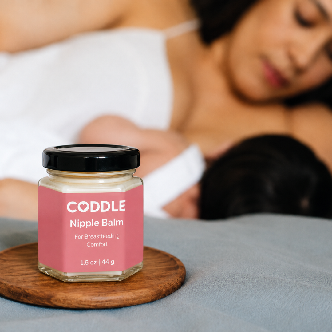 Coddle Nipple Balm sits on a wooden coaster on a bed, with a blurred image of woman in background breastfeeding.