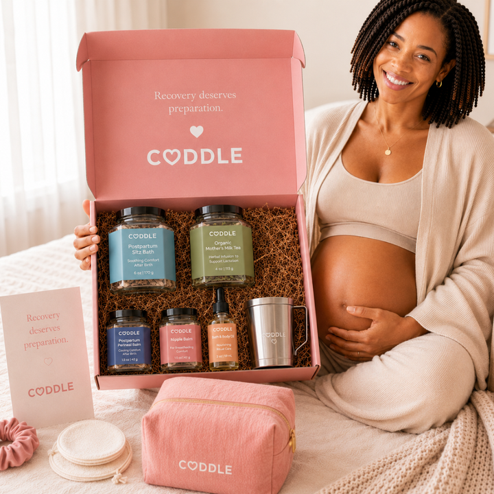 Pregnant woman holding the CODDLE Postpartum Recovery Box featuring Postpartum Sitz Bath, Organic Mother's Milk Tea, Nipple Balm, Postpartum Perineal Balm, Body & Bath Oil, and a stainless steel tea infuser.