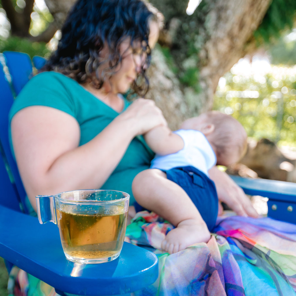 Breastfeeding mother holding baby with a cup of herbal lactation tea for postpartum milk supply support