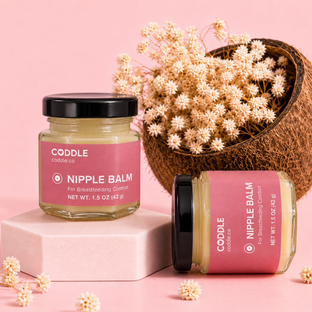 Two CODDLE Nipple Balm hexagonal glass jars with premium rose labels displayed on a blush studio backdrop with dried flowers and a coconut shell, formulated to provide breastfeeding comfort.