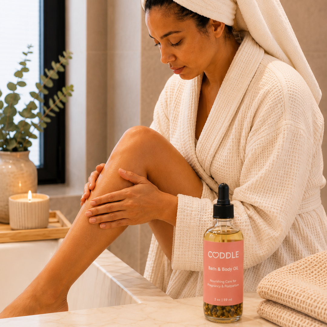 Mother moisturizing her leg with CODDLE Bath & Body Oil in a warm spa-inspired bathroom, featuring a cream waffle robe and nourishing body oil for pregnancy and postpartum care.