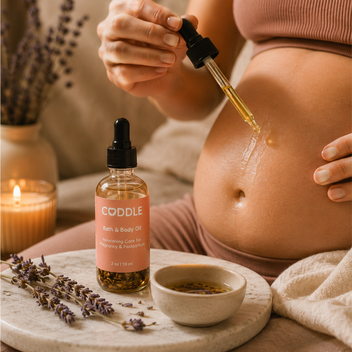 Pregnant mother using CODDLE Bath & Body Oil during a calming self-care ritual, applying nourishing pregnancy and postpartum body oil with a dropper in a warm spa-inspired setting.