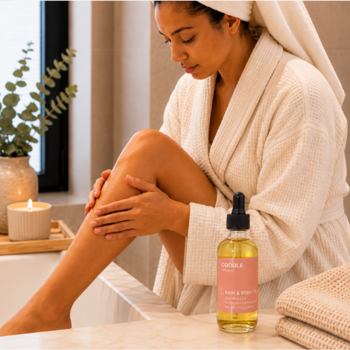 Mother moisturizing her leg with CODDLE Bath & Body Oil in a warm spa-inspired bathroom, featuring a cream waffle robe and nourishing body oil for pregnancy and postpartum care.