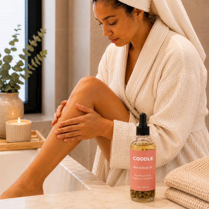Mother moisturizing her leg with CODDLE Bath & Body Oil in a warm spa-inspired bathroom, featuring a cream waffle robe and nourishing body oil for pregnancy and postpartum care.
