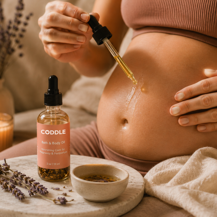 Pregnant mother using CODDLE Bath & Body Oil during a calming self-care ritual, applying nourishing pregnancy and postpartum body oil with a dropper in a warm spa-inspired setting.