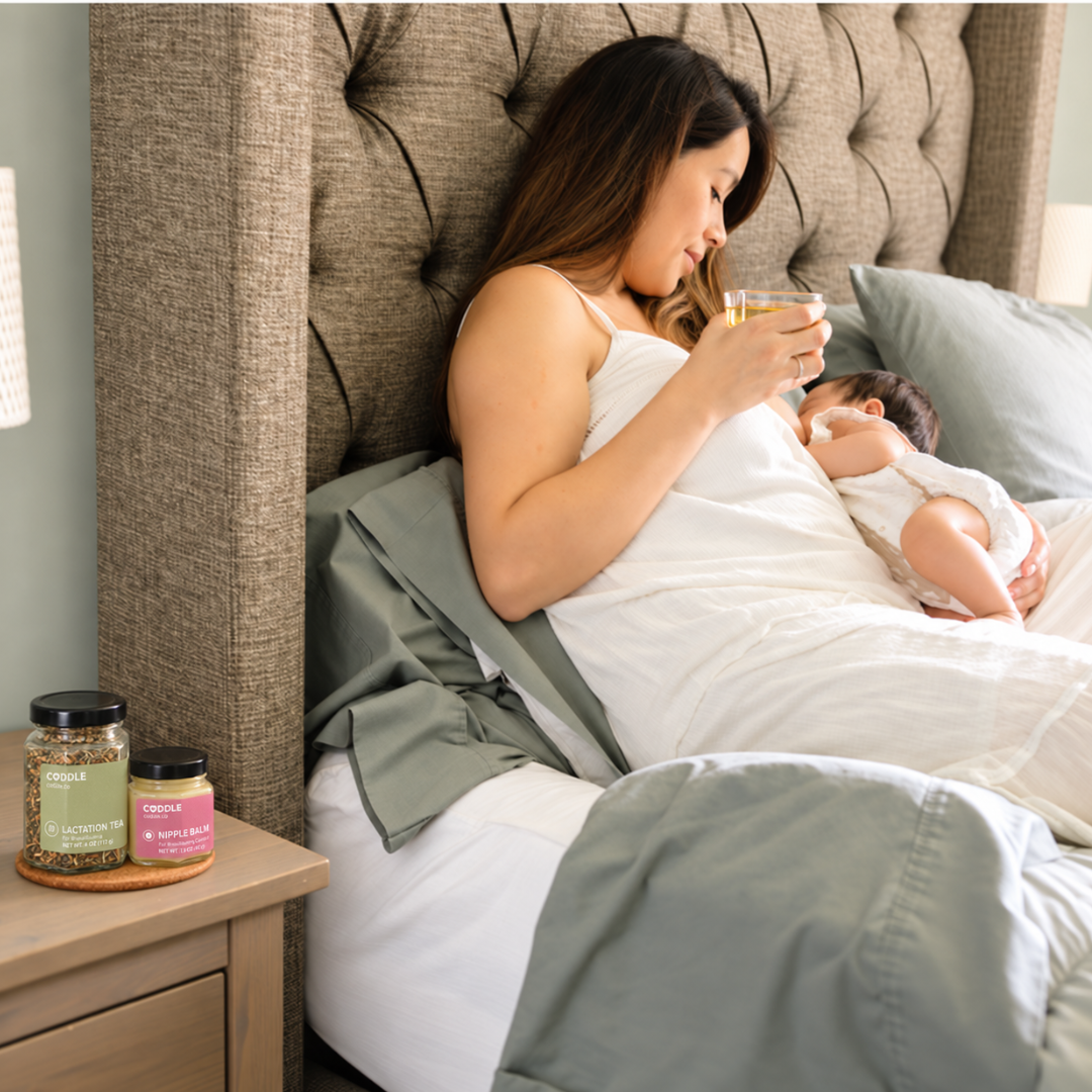 Mother breastfeeding her baby in bed while holding a cup of tea, with CODDLE Lactation Tea and Nipple Balm on the bedside table.