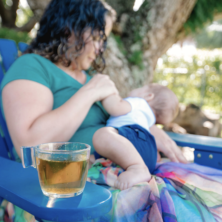 Mother breastfeeding her baby outdoors with a warm cup of CODDLE herbal lactation tea beside her.