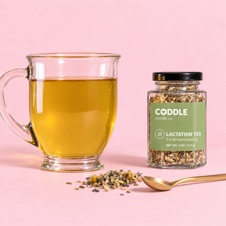 CODDLE Lactation Tea beside a glass cup of prepared herbal tea with loose herbs and a gold spoon on a blush-pink background.