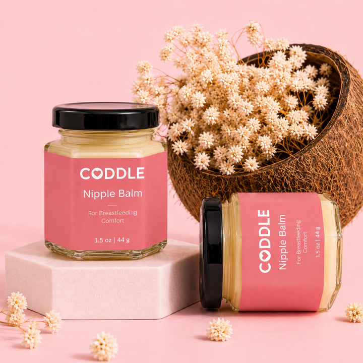 Two CODDLE Nipple Balm hexagonal glass jars with premium rose labels displayed on a blush studio backdrop with dried flowers and a coconut shell, formulated to provide breastfeeding comfort.