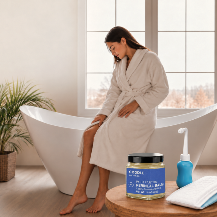 Woman in a cream robe beside a bathtub, with Coddle Postpartum Perineal Balm, a peri bottle and pads on a wooden table.