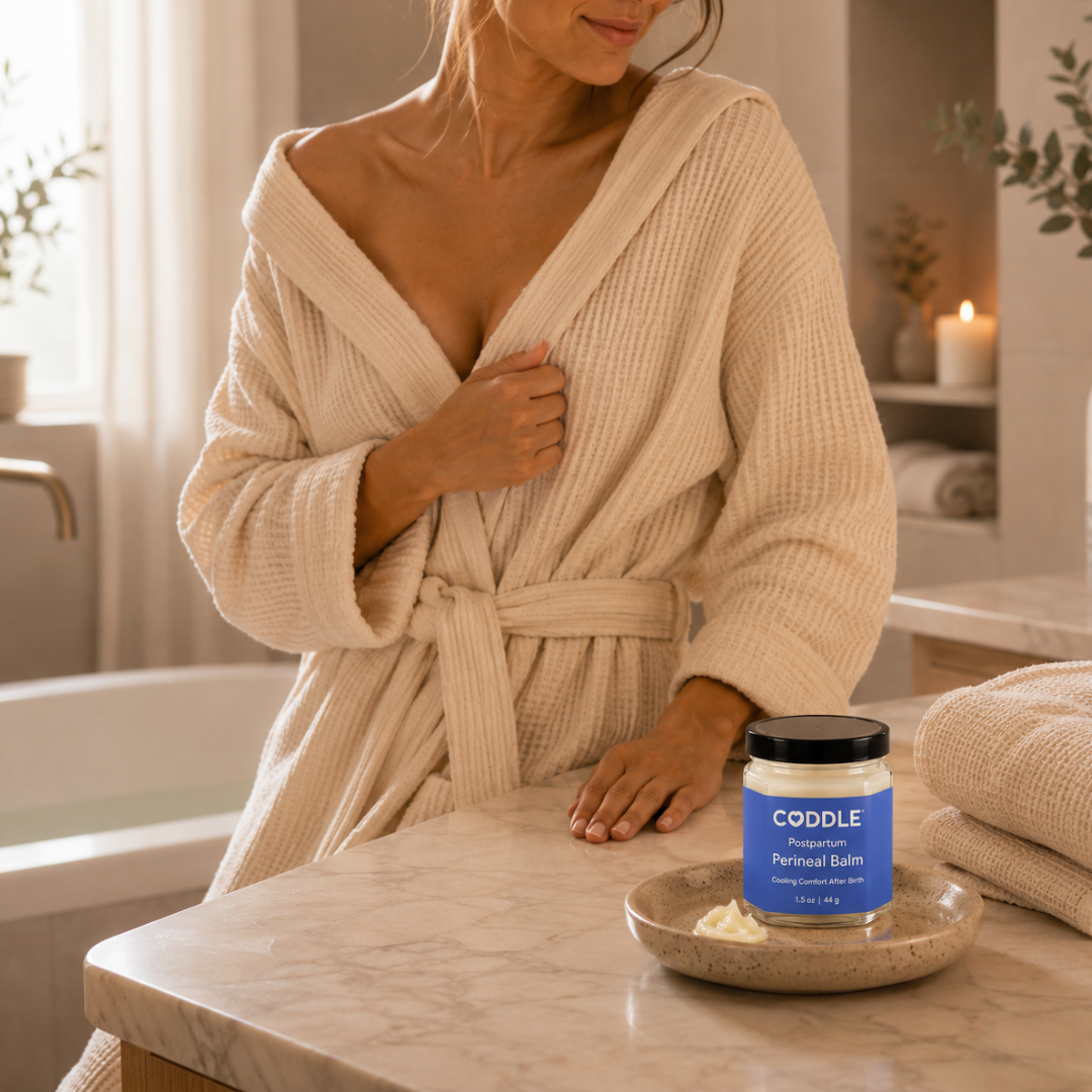 Mother in a cream waffle robe after a sitz bath with CODDLE Postpartum Perineal Balm displayed on a marble bathroom counter, creating a calming postpartum recovery ritual.