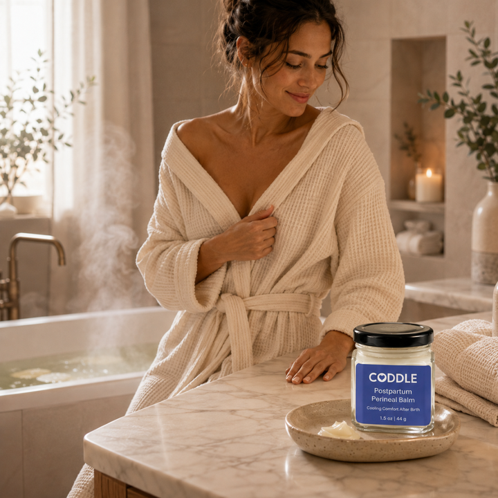 Mother in a cream waffle robe after a sitz bath with CODDLE Postpartum Perineal Balm displayed on a marble bathroom counter, creating a calming postpartum recovery ritual.