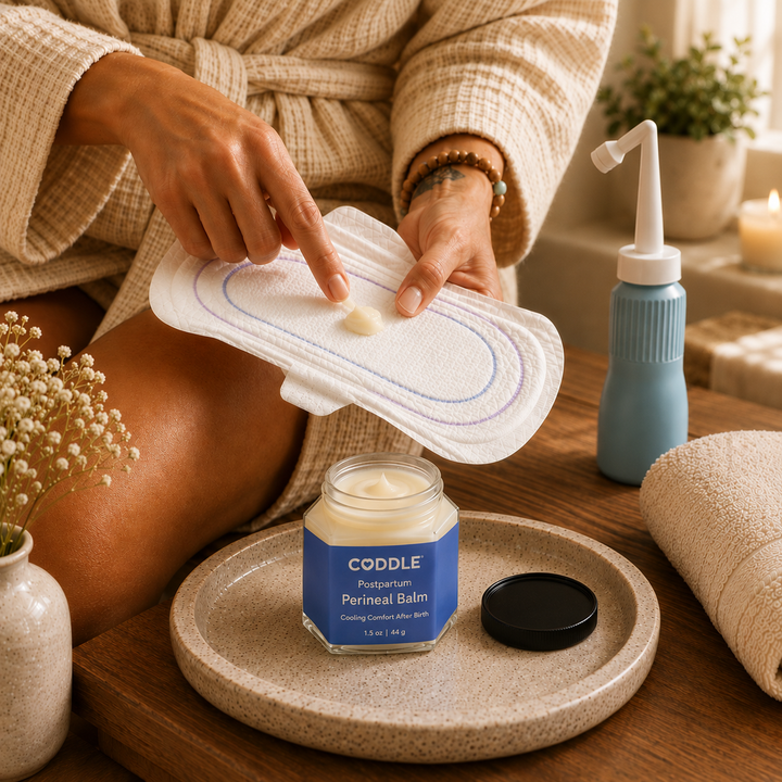 Woman in a cream waffle robe preparing a postpartum cooling pad with CODDLE Postpartum Perineal Balm beside a peri bottle and folded towel, demonstrating a calming postpartum recovery ritual.