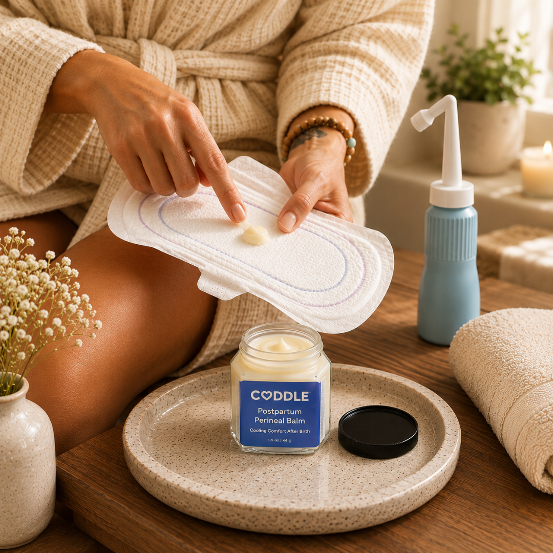 Woman in a cream waffle robe preparing a postpartum cooling pad with CODDLE Postpartum Perineal Balm beside a peri bottle and folded towel, demonstrating a calming postpartum recovery ritual.