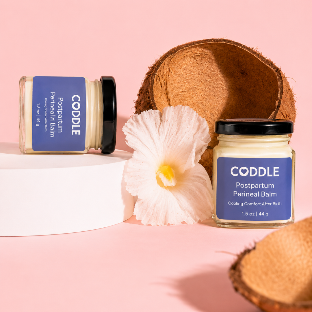 Two CODDLE Postpartum Perineal Balm hexagonal glass jars with blue labels displayed on a blush pink background with coconut shells and a white flower, highlighting cooling comfort after birth.