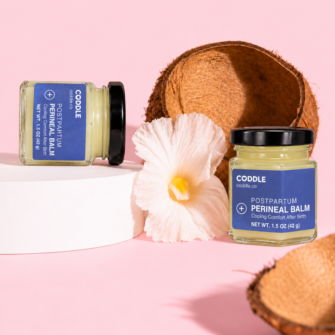 Two CODDLE Postpartum Perineal Balm hexagonal glass jars with blue labels displayed on a blush pink background with coconut shells and a white flower, highlighting cooling comfort after birth.