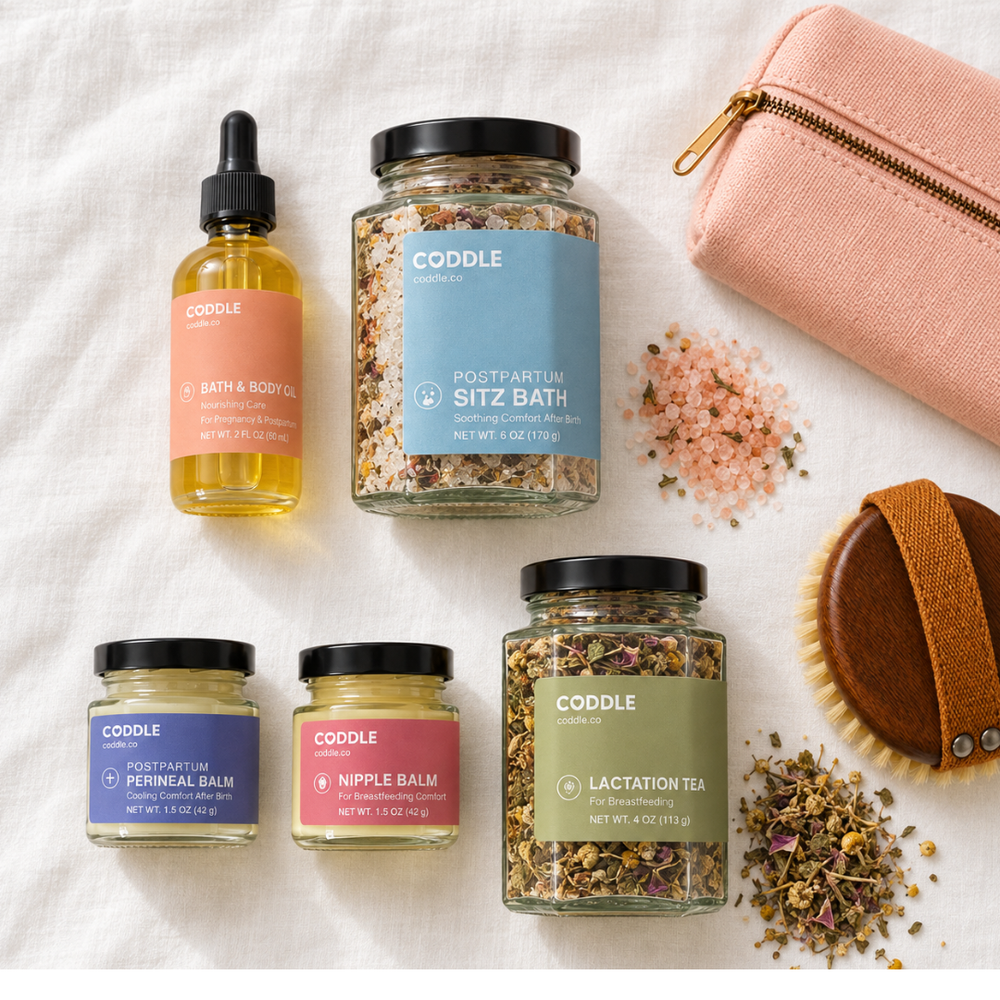 Flat lay of the CODDLE Postpartum Recovery Kit featuring Postpartum Sitz Bath, Bath & Body Oil, Postpartum Perineal Balm, Nipple Balm, and Organic Mother's Milk Tea arranged on a white linen background with postpartum recovery essentials.