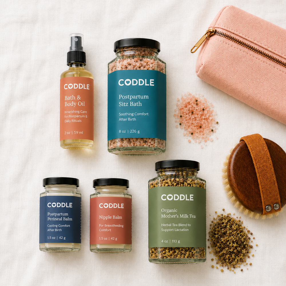 Flat lay of the CODDLE Postpartum Recovery Kit featuring Postpartum Sitz Bath, Bath & Body Oil, Postpartum Perineal Balm, Nipple Balm, and Organic Mother's Milk Tea arranged on a white linen background with postpartum recovery essentials.