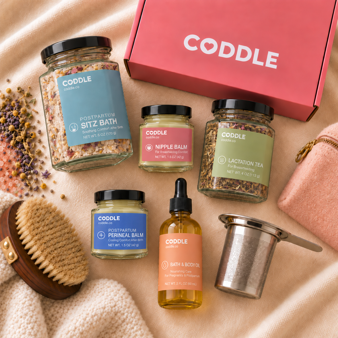 Coddle postpartum recovery products arranged on cream linens beside a pink gift box, body brush and tea infuser.