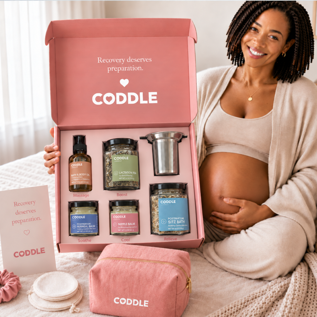 Pregnant woman holding the CODDLE Postpartum Recovery Box featuring Postpartum Sitz Bath, Organic Mother's Milk Tea, Nipple Balm, Postpartum Perineal Balm, Body & Bath Oil, and a stainless steel tea infuser.