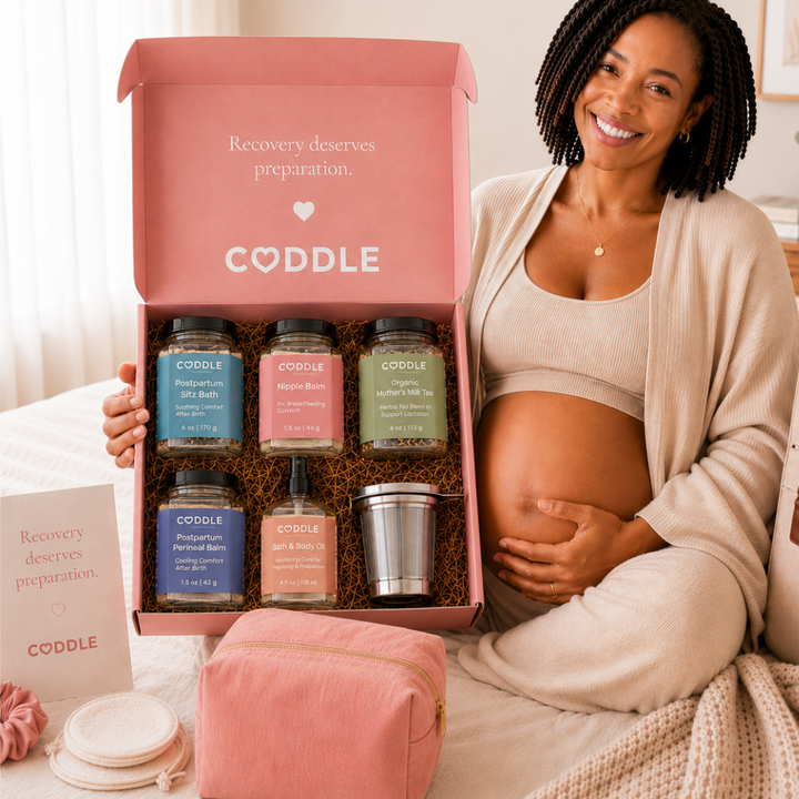 Pregnant woman holding the CODDLE Postpartum Recovery Box featuring Postpartum Sitz Bath, Organic Mother's Milk Tea, Nipple Balm, Postpartum Perineal Balm, Body & Bath Oil, and a stainless steel tea infuser.