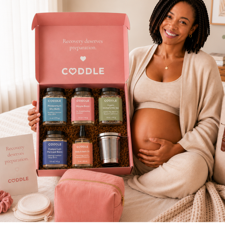 Pregnant woman holding the CODDLE Postpartum Recovery Box featuring Postpartum Sitz Bath, Organic Mother's Milk Tea, Nipple Balm, Postpartum Perineal Balm, Body & Bath Oil, and a stainless steel tea infuser.