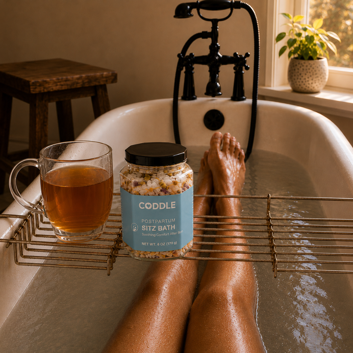 A relaxing postpartum sitz bath with CODDLE Postpartum Sitz Bath herbal blend on a bath caddy beside herbal tea, creating a calming recovery ritual in a warm spa-inspired bathroom.