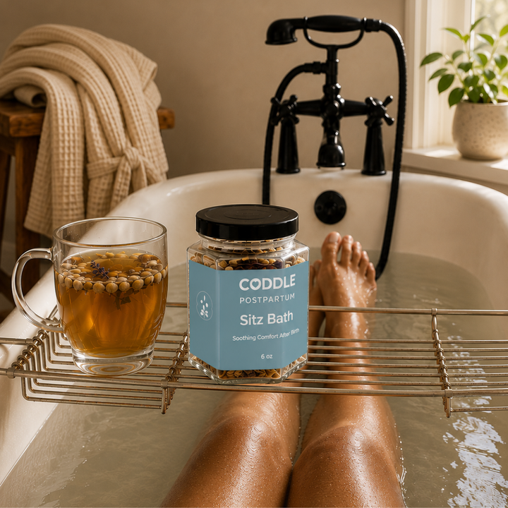View from a relaxing postpartum sitz bath with CODDLE Postpartum Sitz Bath herbal blend on a bath caddy beside herbal tea, creating a calming recovery ritual in a warm spa-inspired bathroom.